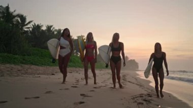 Gün doğumunda sahilde yürüyen sörfçü kızların siluetleri. Sörf tahtaları taşıyan dişi sörfçüler şafak vakti okyanus boyunca sahilde yürüyorlar. Okyanusta yürüyen genç sörfçü kadınlar.