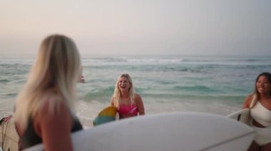 Sörf tahtası tutan mutlu sörfçü kızlar güneşin doğuşunda sahilde neşeyle konuşuyorlar. Gülümseyen dişi sörfçüler şafakta okyanusun kenarında dururlar. Deniz kenarında sörf yapan genç kadınlar ve dalgalar gülüyor ve denize bakıyor.