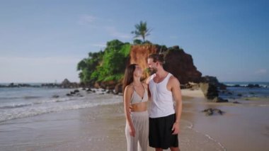 Video portrait of happy diverse couple embracing and smiling to camera at exotic tropical seaside resort against picturesque rock on background. Cheerful biethnic couple looking at camera. Inclusivity