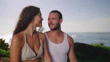 Young multiracial couple sitting at seaside huhhing, smiling and looking aside pointing at something frontview shot. Happy biethnic couple at scenic background embracing talking cheerfully closeup