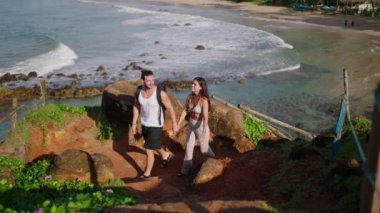 Biracial tourist couple with backpacks walking up scenic rocky hill covered with greenery at exotic spot pointing at something and enjoying the view. Young adults climbing picturescue rock down shot