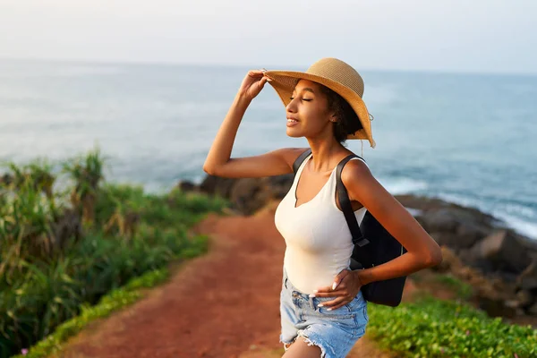 Stock image Multiethnic woman in straw hat enjoys tropical vacation travel holiday. Black female with backpack sightseeing on scenic location. Pretty lady standing on island on sunset. Girl in the sun for suntan