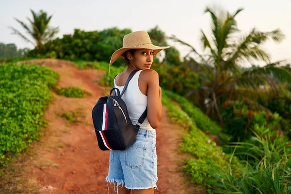 stock image Multiethnic woman in straw hat enjoys tropical vacation travel holiday. Black female with backpack sightseeing on scenic location. Pretty lady standing on island on sunset. Girl in the sun for suntan