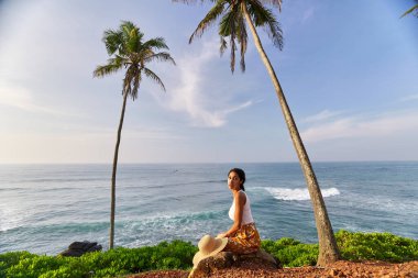 Young african female model posing in colorful clothes at scenic tropical location by ocean between palms at sunrise. Black woman sitting against exotic scenery with view of sea and palm trees at dawn