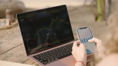 Akıllı telefondan istatistik çizelgelerini kontrol eden bilgisayardaki kadın kripto tüccarı. Şifreli para birimiyle uzaktan çalışan bir kadın telefonda şema kontrol ediyor. Şifreleme grafiklerini yakından inceleyen kadın analist