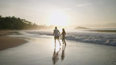 Silhouettes of young happy couple holding hands and running on the beach together enjoying summer back view. Boyfriend and girlfriend relax and walk at the seaside hugging and kissing at sunrise