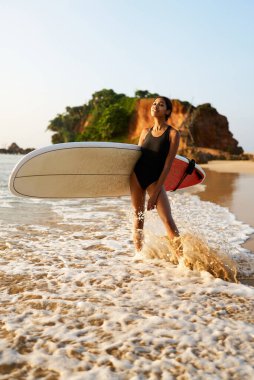 Okyanus sahilinde sörf tahtası olan Afrikalı Amerikalı bir kadın. Siyah sörf tahtası ile poz veren kadın sörfçü. Çok ırklı bir kız güneş doğarken tropikal bir yerde sörf yapmaya gidiyor..