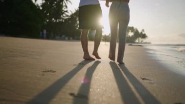 Legs of happy couple holding hands and walking on the beach together enjoying summer back view. Feet of heerful boyfriend and girlfriend relaxing and taking walk at the seaside at sunrise back shot.