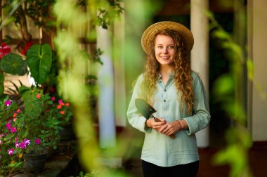 Bahçede elinde bahçe aletleriyle duran genç bir kadın gülümseyerek kameraya bakıyor. Yeşil meyve bahçesinde poz veren genç bayan bahçıvan. Çiftçilik ve bahçecilik kavramı.