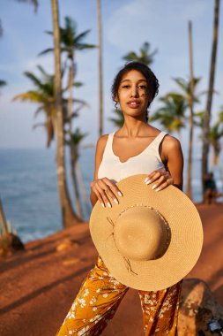 Young african female model posing in colorful clothes at tropical setting at sunrise. Black woman against exotic scenery at dawn. Multiracial dark-skinned model poses in front of palm trees at sunset