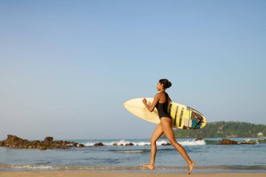 Okyanus sahilinde sörf tahtasıyla koşan Afrikalı Amerikalı bir kadın. Sörf tahtası olan siyah bir kadın sörfçü. Çok ırklı bir kız güneş doğarken tropikal bir yerde sörf yapmaya gidiyor..