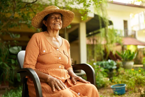 Happy senior Indian woman sitting in chair in garden of her house smiling looking at camera. Elderly retired Sri Lankan cheerful female in green orchard at her villa with plants in the background
