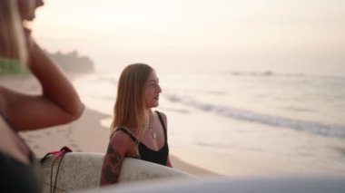 Güneşin doğuşunda sörf tahtası tutan gülümseyen sörfçü kız arkadaşlarıyla gülüp konuşuyor. Neşeli dişi sörfçüler şafakta okyanusun kenarında dururlar. Genç sörfçü kadınlar eğlenmek için yer arar.