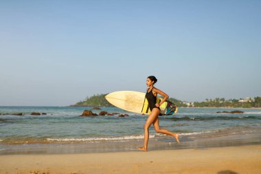 Okyanus sahilinde sörf tahtasıyla koşan Afrikalı Amerikalı bir kadın. Sörf tahtası olan siyah bir kadın sörfçü. Çok ırklı bir kız güneş doğarken tropikal bir yerde sörf yapmaya gidiyor..