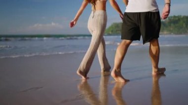 Legs of happy couple holding hands and walking on the beach together enjoying summer back view. Feet of heerful boyfriend and girlfriend relaxing and taking walk at the seaside at sunrise back shot.