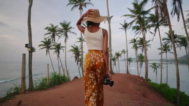 アフリカ系アメリカ人の女性が熱帯の島でカメラを持って歩く 女性の多人種写真家観光客は休暇中に海の景色の場所を探索します 黒女の子でわら帽子撮影写真上の日の出 — ストック動画
