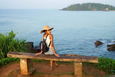 Hasır şapkalı çok ırklı kadın, deniz manzaralı kayalık kayalıklarda oturarak tropikal tatilin tadını çıkarıyor. Sırt çantalı siyah bir kadın manzaralı yerleri geziyor. Kız okyanus batımında adada bronzlaşıyor..