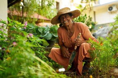 Kıdemli Hintli kadın elinde bahçe kepçesi tebessümleri meyve bahçesinde oturmuş yere çiçek dikiyor. Yaşlı Sri Lankalı mutlu kadın bahçede bitkilerle ilgileniyor. Bahçe işleri konsepti. Mutlu emeklilik