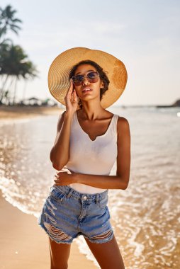 Young african female model in a straw hat and sunglasses posing at resort by sea at sunrise. Black woman against scenic rocky green island and ocean tide at dawn. Multiracial model poses on vacation