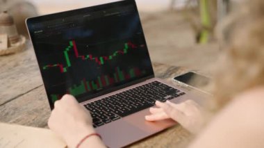 Dizüstü bilgisayardaki kadın kripto para tüccarı Candlestick çizelgelerini kontrol ediyor. Açık hava tropikal deniz kenarı kafesinde çalışıyor. Kadın kripto komisyoncusu hisse senedi kurlarını ve bahisleri okyanusa yakın hesaplıyor.