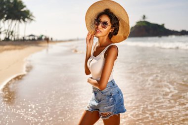 Hasır şapkalı ve güneş gözlüklü genç Afrikalı kadın model güneş doğarken tatil köyünde poz veriyor. Siyah kadın, kayalık yeşil adaya ve şafakta okyanus gelgitine karşı. Çok ırklı model tatilde poz veriyor
