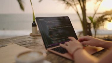 Female developer working on laptop by the ocean. Young woman freelancer coding at outdoor tropical cafe. Caucasian girl works remotely typing on her computer at exotic location. Worldwide work concept