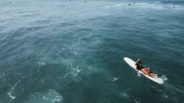 Genç bayan sörfçü güneşli bir günde, hava manzaralı tropik sahilde kristal okyanus suyunda sörf tahtasında kürek çekiyor. Sörfçü kız yaz tatili drone çekimlerinde kürek çekmekten ve sörf yapmaktan hoşlanıyor..