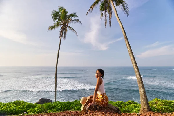 Young african female model posing in colorful clothes at scenic tropical location by ocean between palms at sunrise. Black woman sitting against exotic scenery with view of sea and palm trees at dawn