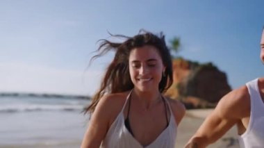 Young happy couple runs holding hands on beach through waves splashing water and enjoying summer closeup shot. Boyfriend and girlfriend having fun at spectacular scenic seaside tropical location.