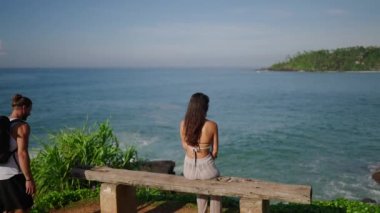 Guy approaches girl sitting alone on bench in front of sea and hugs cinematic shot. Man comes to bench by ocean with woman sitting, embraces her. Couple on bench at the edge of scenic hill back shot