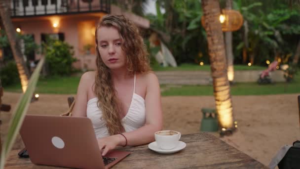 在户外咖啡馆里有笔记本电脑的女性在度假胜地喝咖啡并开始工作 在热带地区的计算机上打字的白人妇女自由职业者从侍者那里得到了咖啡 她微笑着继续工作 — 图库视频影像