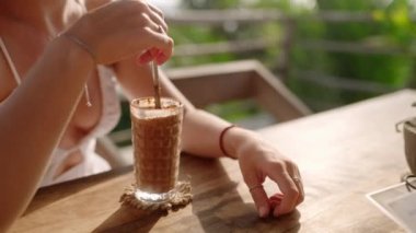 Kadın elleri açık hava restoranında metal pipetle kokteyl karıştırıyor. Genç bir bayan güneşli tropikal kafede çikolatalı milkshake içiyor. Masadaki kız büyük bardakta buzlu kahve içiyor.