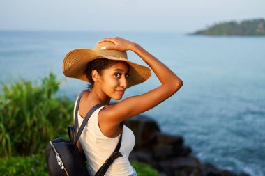 Hasır şapkalı çok ırklı kadın, deniz manzaralı kayalık kayalıklarda oturarak tropikal tatilin tadını çıkarıyor. Sırt çantalı siyah bir kadın manzaralı yerleri geziyor. Kız okyanus batımında adada bronzlaşıyor..