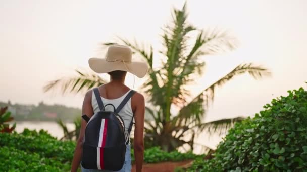 アフリカ系アメリカ人の女性が熱帯の島のバックパックでハイキング 藁帽子の多人種の女性が海を歩いています 黒の可愛い女の子は海岸を探検する バックビュー 旅行休暇中の観光 — ストック動画