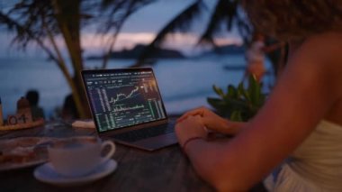 Bilgisayardaki kadın kripto para tüccarı günbatımında açık deniz kafesinde çalışan online haritaları kontrol ediyor. Kadın kripto komisyoncusu okyanusa yakın plandaki borsa oranlarının grafiklerini analiz ediyor.