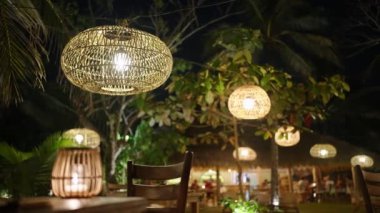 Geceleri ağaçlardan sarkan hasır lambaları tropikal arka planda süsleme olarak kullanılıyor. Dışarıdaki kafede eski moda ahşap ampuller sallanıyor. Egzotik ortamda Boho düğün süslemeleri.