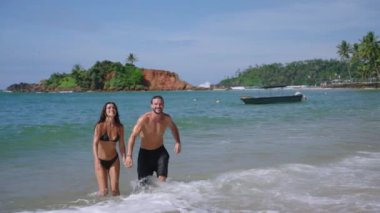 Young cheerful biracial couple in swimsuits holding hands running out of ocean waves splashing them slow motion shot. Multiracial happy man and woman run joyfully out of sea in slowmo front view