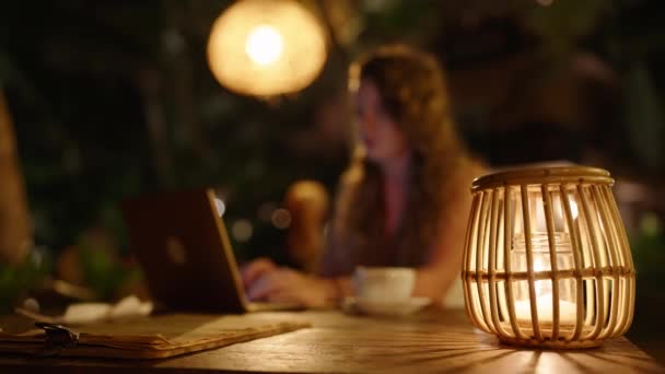 女性のフリーランスは 大気圏外の熱帯地域で夜に働いています 暖かい照明とわらとノートパソコンを使用して木製のランプと暗いカフェの外のテーブルに座っている女性 遅くまで働く — ストック動画