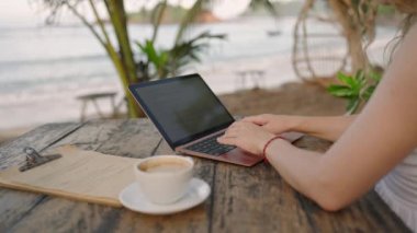 Young woman working on laptop sitting in outdoor cafe in nature. Female freelancer typing on computer at tropical location. Young adult works remotely at exotic location checks the menu chooses food