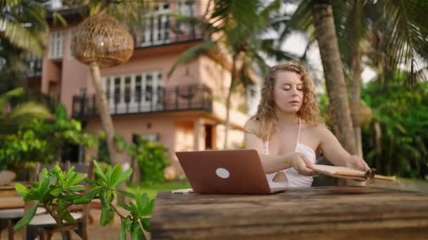 白人妇女坐在度假胜地的室外咖啡馆里 带着笔记本电脑 在热带地区的计算机上打字的女自由职业者 在异国他乡工作的年轻人检查选择食物的菜单 — 图库视频影像