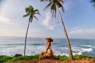 Young african female model posing in colorful clothes at scenic tropical location by ocean between palms at sunrise. Black woman sitting against exotic scenery with view of sea and palm trees at dawn