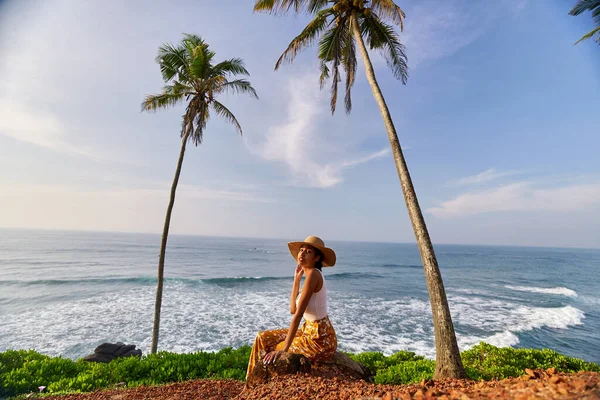 Young african female model posing in colorful clothes at scenic tropical location by ocean between palms at sunrise. Black woman sitting against exotic scenery with view of sea and palm trees at dawn