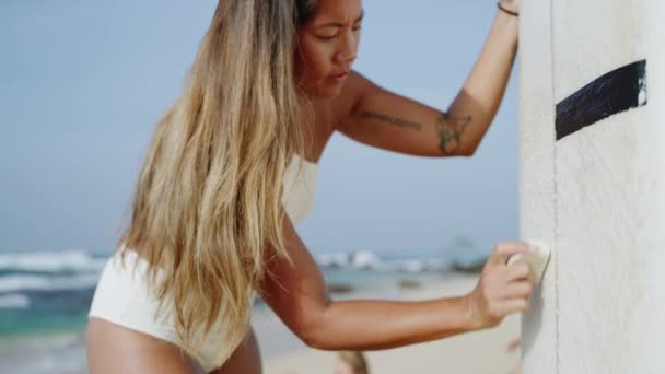 亚洲冲浪女孩在海滩的冲浪板上擦蜡 为会议做准备 穿着白色泳衣的晒黑了的Mestizo女冲浪手在海边给她的冲浪板上蜡 年轻女人用蜡盖板 — 图库视频影像