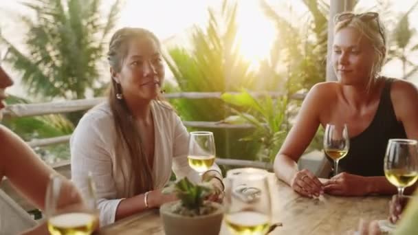 多様な女性の友人が日没時に屋上のオーシャンビューレストランでワインを飲みます 多人種の女性が話し 海沿いのトロピカルカフェで笑顔 アジア系 インド系 白人系の若い女性が食事をする — ストック動画