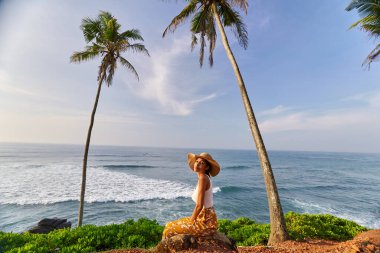 Young african female model posing in colorful clothes at scenic tropical location by ocean between palms at sunrise. Black woman sitting against exotic scenery with view of sea and palm trees at dawn