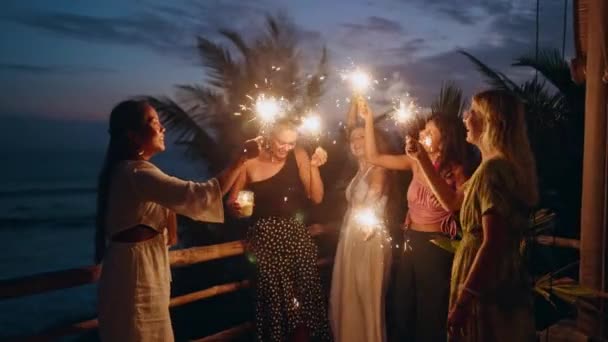 若い多人種の女性は輝くライトと海で熱帯のクリスマスを祝う楽しみを持っています 多様な幸せな女性が海辺の独立記念日パーティーで輝く人々を応援し踊る — ストック動画