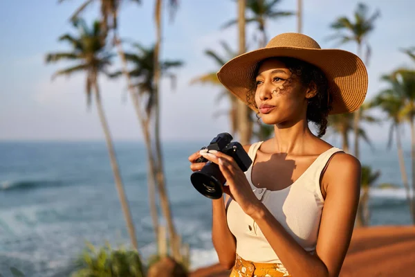 Young african female model tourist with camera in colorful clothes and straw hat taking pictures at tropical location at sunrise. Black travelling woman takes photos in exotic ocean scenery at dawn