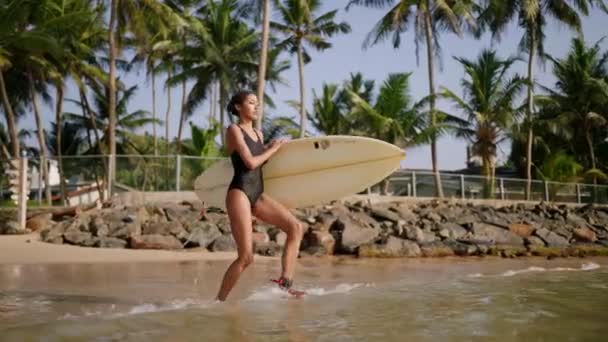 アフリカ系アメリカ人の女性が海の水にサーフボードで走り 漕いでいる サーフボード付きの黒い女性サーファー かなり多民族の女の子行きますサーフィンセッションに熱帯の場所で日当たりの良い — ストック動画