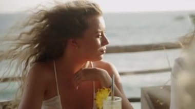 Deniz manzaralı beyaz elbiseli sarışın genç bayan konuşuyor, gülümsüyor, rüzgar uzun saçlarını savuruyor. Gün batımında deniz kenarındaki bir restoranda genç ve mutlu bir kıvırcık kadın kızlarla sohbet ediyor. Okyanus görünümü çubuğu