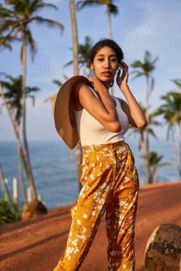 Young african female model posing in colorful clothes at tropical setting at sunrise. Black woman against exotic scenery at dawn. Multiracial dark-skinned model poses in front of palm trees at sunset